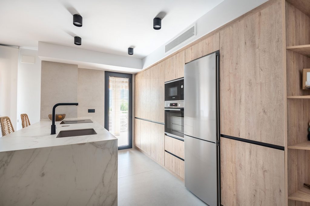 Diseño de cocina a medida con baldas de madera natural rechapada para dar continuidad y prolongar el espacio entre la cocina y el comedor.